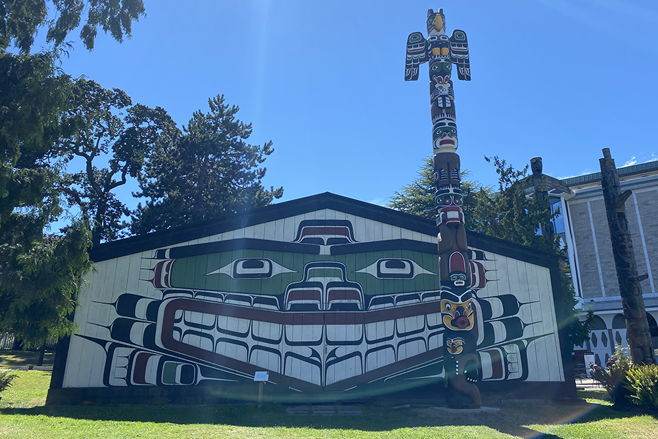Grassy lawn, bright sun and a ceremonial big house with totem poles against a backdrop of evergreen trees.