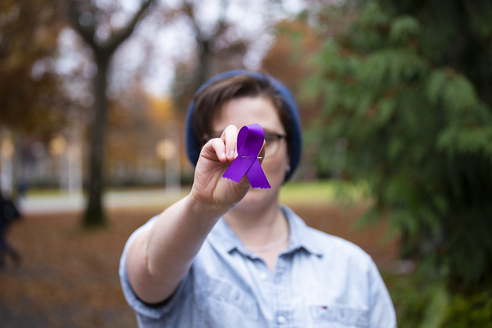 Close-up of person holding purple ribbon to camera, UVic campus in background