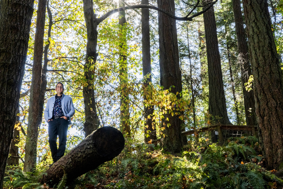 Wide-angle photo of Ian Mauro standing on a log with hands in pockets in forested setting
