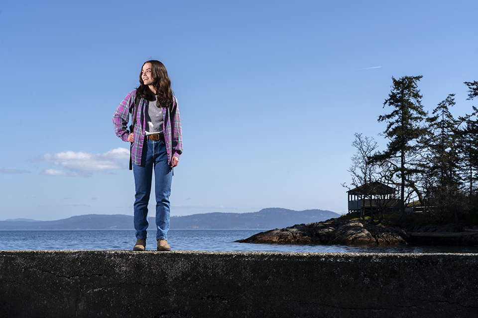 Smiling university student stands on a cement wall that overlooks the ocean and they look to their right.