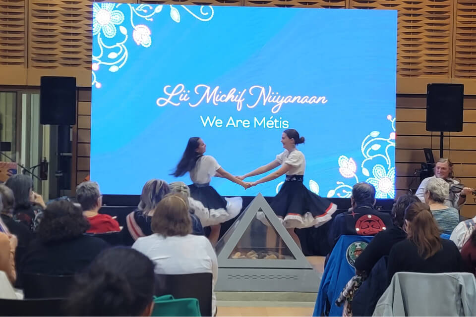 Metis jiggers holding hands and dancing infront of the LED screen backdrop. Credit: Hannah Mashon