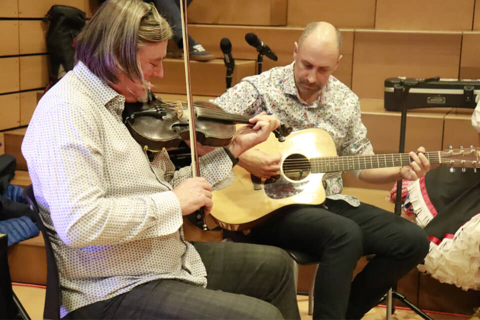 Two male musicians sitting beside each other playing a fiddle and an acoustic guitar.