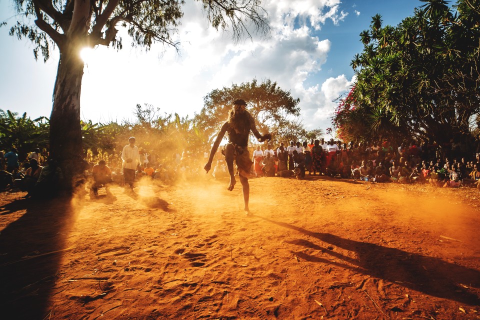 A masked person dances in the dust amongst a group of onlookers in southern Africa.