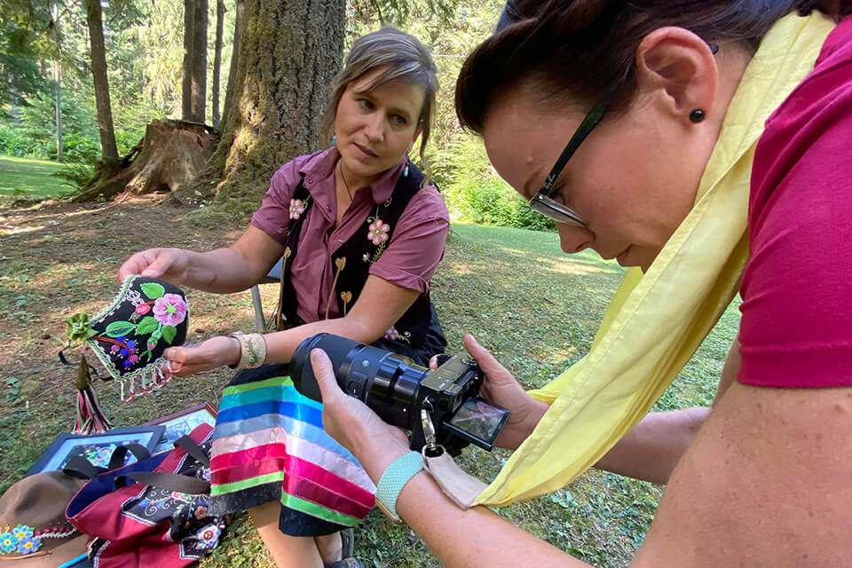 Métis beadwork artist Lisa Shepherd with a beaded piece of artwork with film director Madeline Ell holding a camera for a close-up shot in a forest.