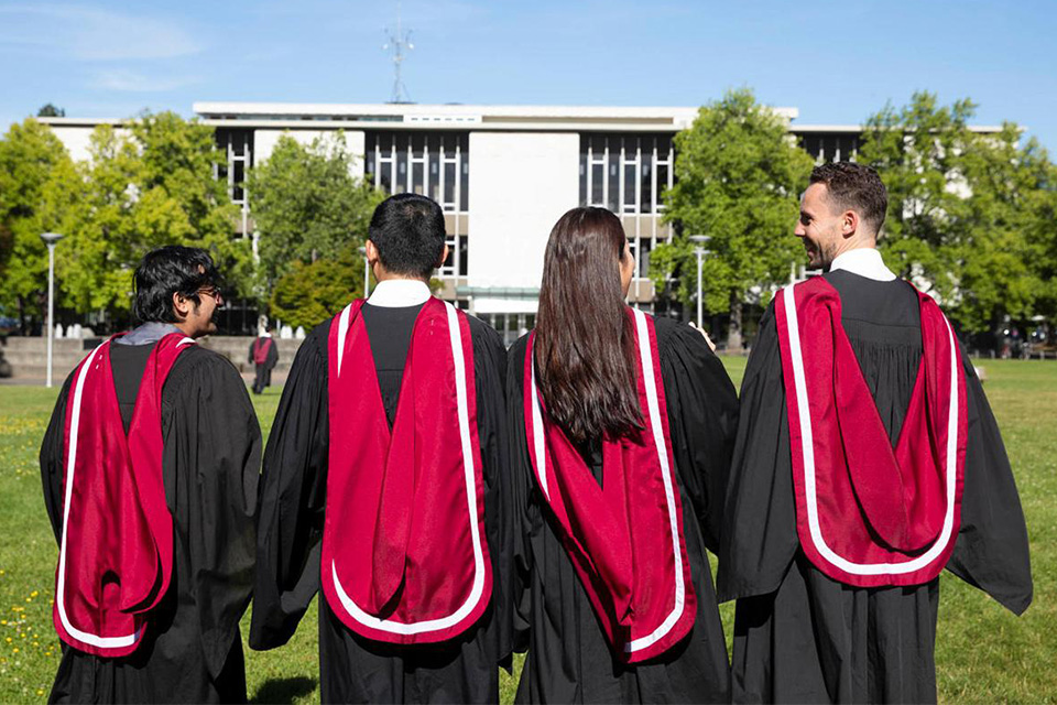 The backs of four graduating students wearing black gowns and red regalia hoods walking outside towards a building.