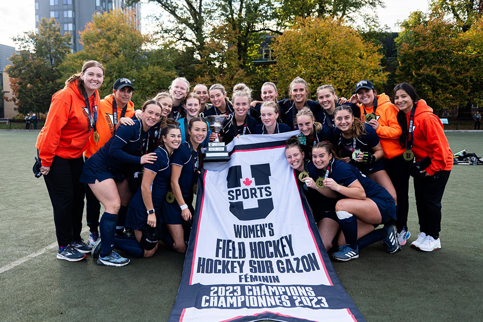 The University of Victoria women's field hockey team celebrates their fifth U SPORTS Championship by posing with the banner and their gold medals. 