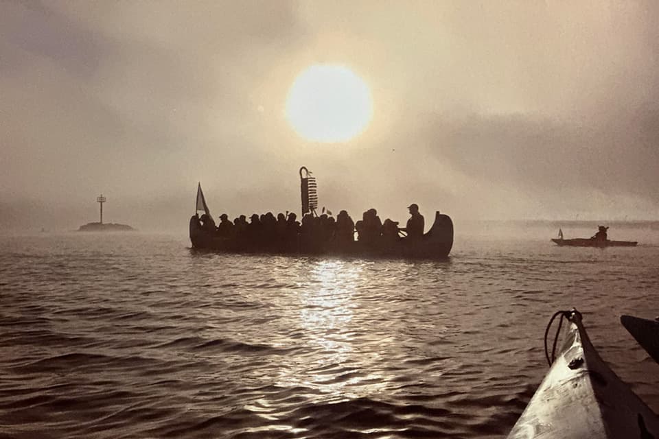 A full Indigenous cedar dugout canoe on the water in slightly foggy conditions. Morning sun rising in the background. 