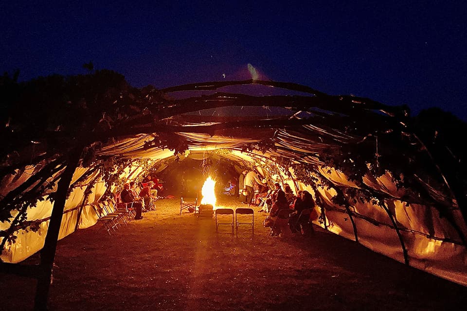 Looking into a large teaching lodge structure at night, curved walls and ceiling made of cloth and wooden poles. Open at each end. a large group of people sit on either side around a fire in the middle.