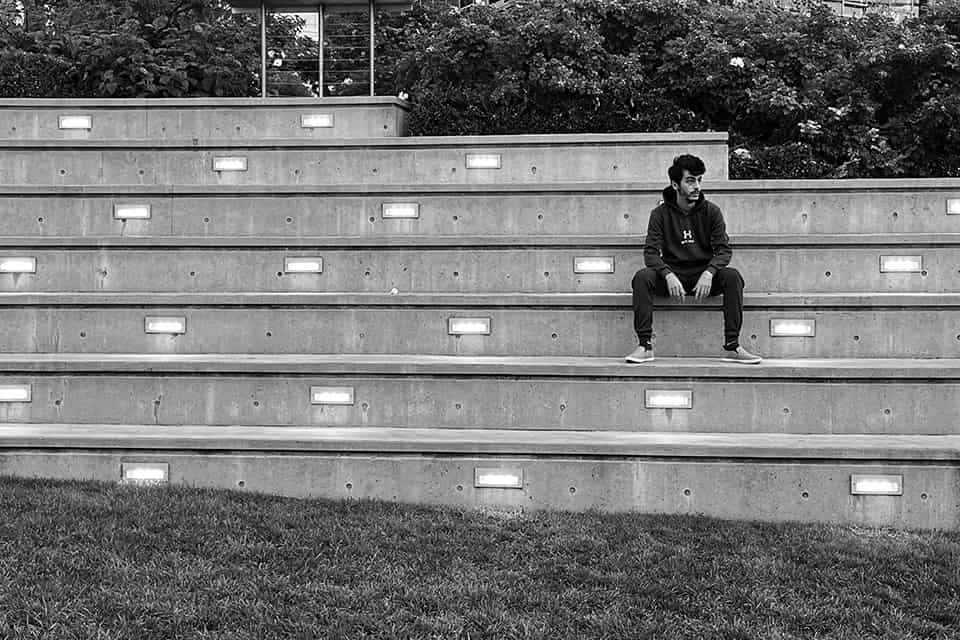 black and white photo of a man in a dark hooding and track pants sitting alone on 7-tiered concrete bleacher