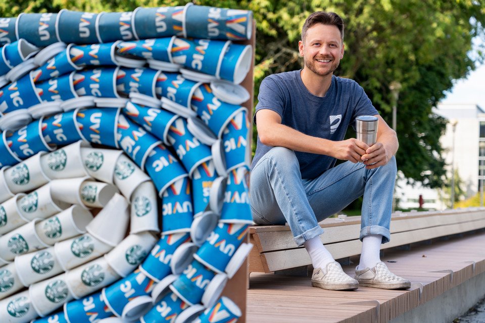 Matt Miller, ​UVic PhD student ⁣in Oceanography, behind a collage of‍ UVic single-use beverage ⁣cups.