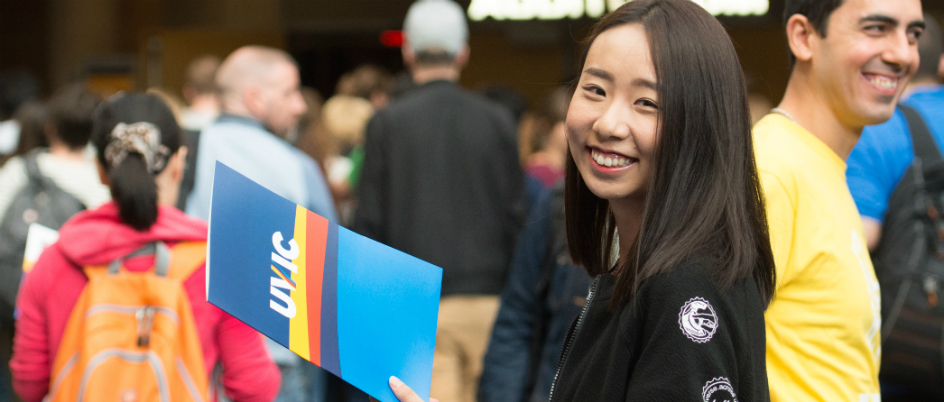 Female student smiling at the camera holding a UVic folder, in front of a blurred crowd entering an auditorium