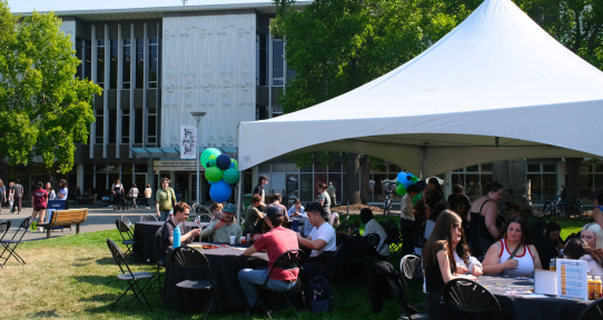 Students talking and playing games at tables under a tent on the quad