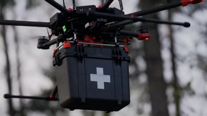 A drone carries a black box marked with a white cross indicating medical supplies