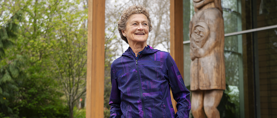 A photo of Cecilia Benoit in a purple zipped sweater standing in front of the First People's House at UVic with a totem pole in the background.