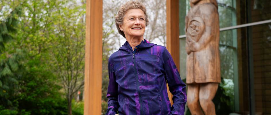 Cecilia Benoit, dressed in a purple sweater, stands in front of First Peoples House at the University of Victoria. A totem pole and greenery are in the background.