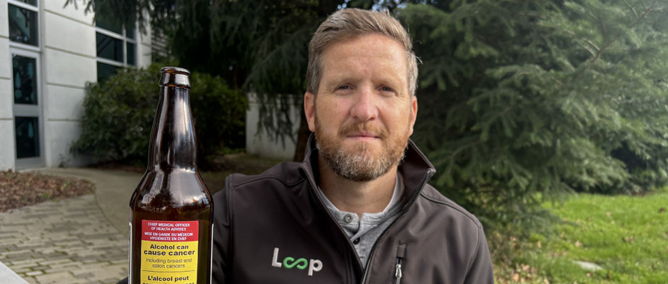 A photo of CISUR scientist Adam Sherk sitting at a picnic table holds a beer bottle with a mock-up of a cancer warning label. He is lead author of a new study looking at the impact cancer warning labels and pricing tied to alcohol content could have on cancer cases and deaths in Canada.