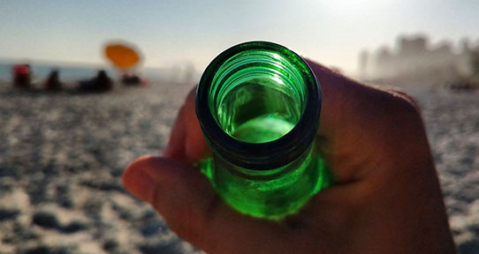 A photo of the mouth of a green glass bottle being tilted towards the camera. A beach is in the background.
