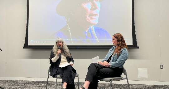 Joan Johnson (left) and Andrea Cowan at the Harold Johnson Award. They are sitting in front of a video screen with a video of Harold Johnson in the background.