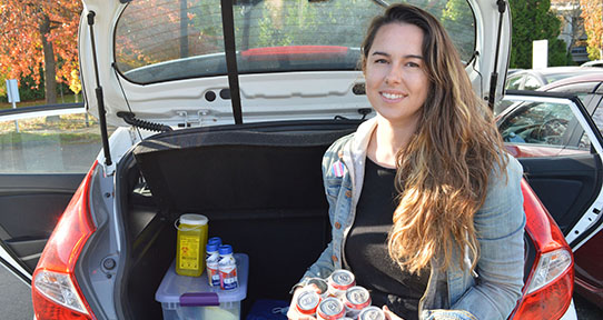 A photo of a woman sitting in the trunk of a car, holding a six pack of beer. There is a bin of PPE, a sharps container and ensures beside her.