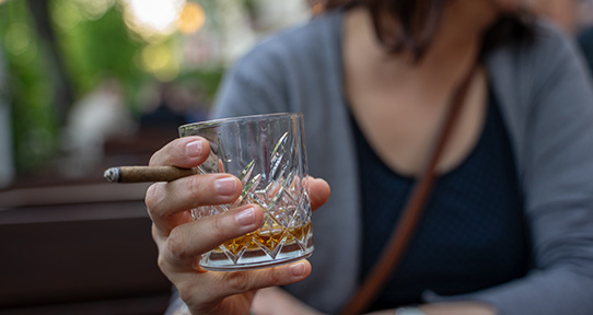 A photo of someone holding a small tumbler of whiskey and a small cigar.