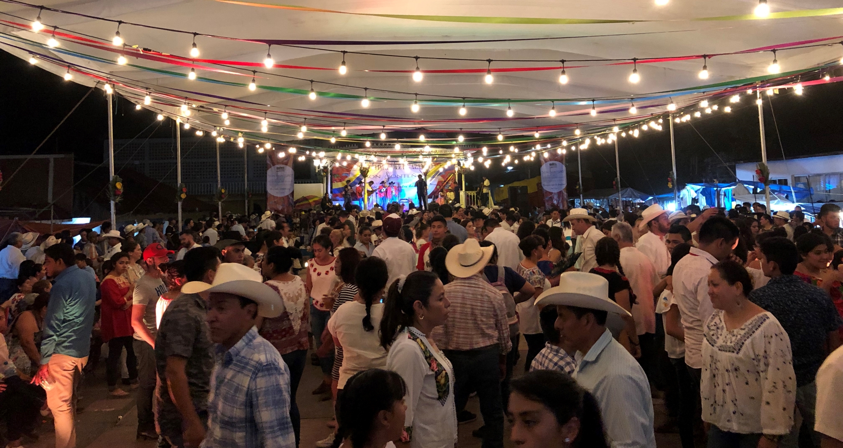A large group of people interacting under string lights and colourful streamers. A band plays in the background. 
