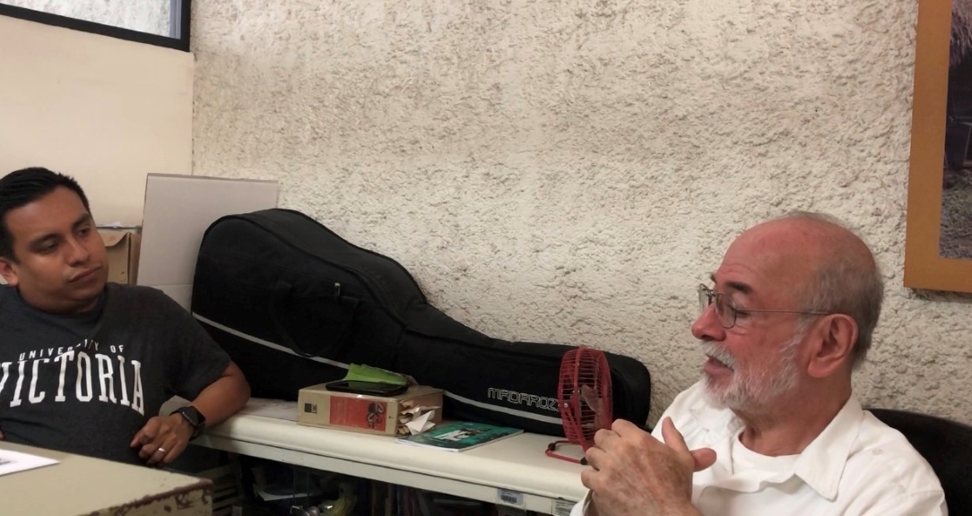 Héctor sits opposite an older man. They appear to be deep in conversation.