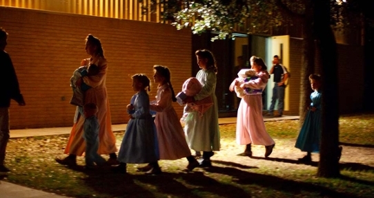 A group of young women and girls in dresses carrying piles of belongings outside a church at night