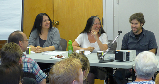 A photo of two woman and a man sitting at a table speaking to a crowd. Woman in the middle is laughing.
