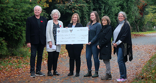 A photo of six people holding an oversized cheque while standing on a paved path. Chevalier Dr. Richard Crow (far left), Dame the Honorable Mary Collins, Dr. Kelli Stajduhar (UVic), Robyn Kyle (PORT, Island Health), Maria Dominelli (Former Commander), Carren Dujela (UVic)