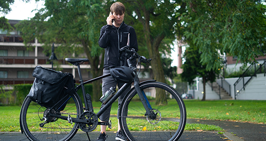 A photo of a woman in a black hoodie and pants standing behind a black bicycle. She is talking in a cellphone.