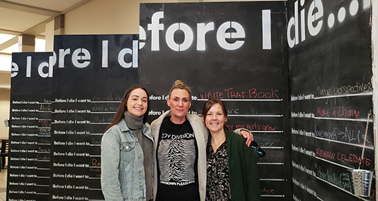 Three women stand in front of a multiple blackboards with "before I die..." written at the top