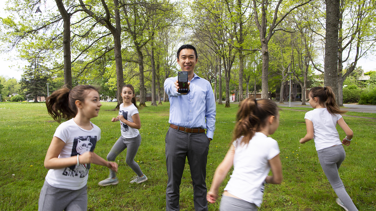 UVic researcher Sam Liu stands holding phone on the quad with kids running around him