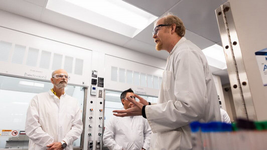 David Leitch in a lab coat and goggles speaking to two other men in a lab