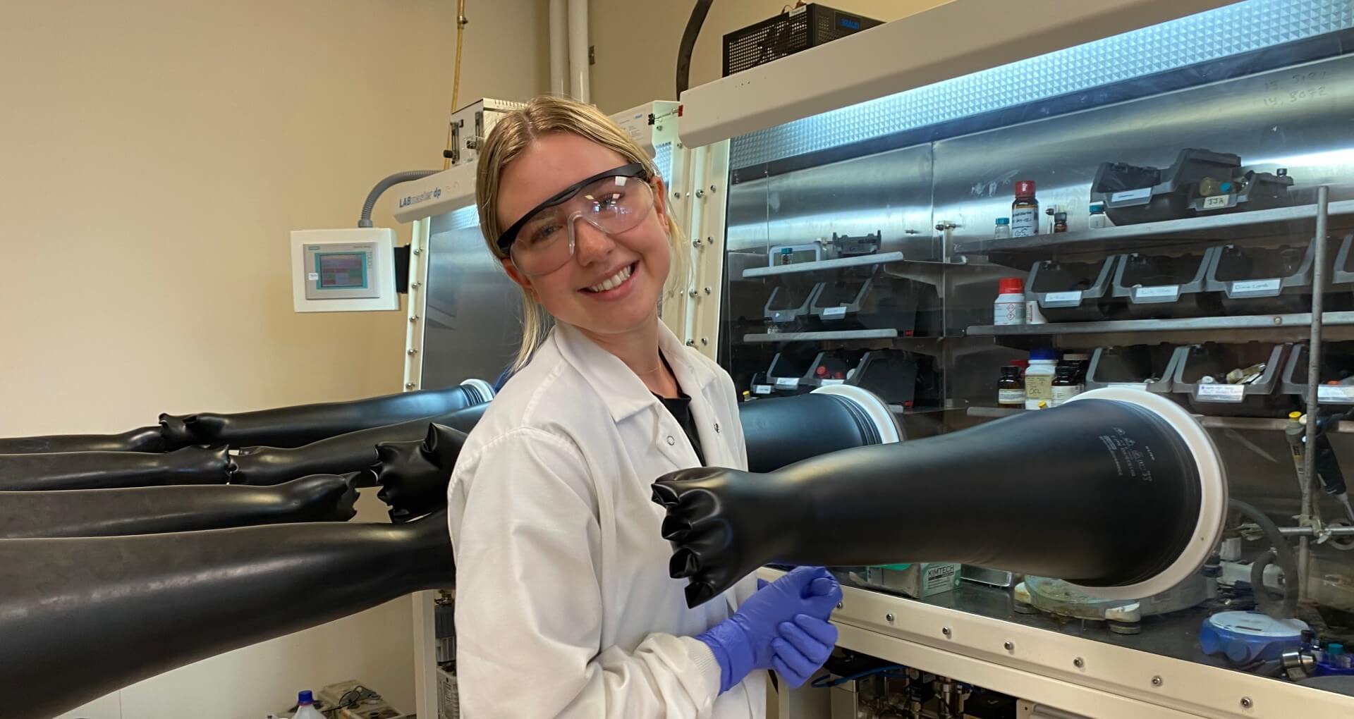 Hailey Gray in a lab coat and gloves standing in front of a glove box