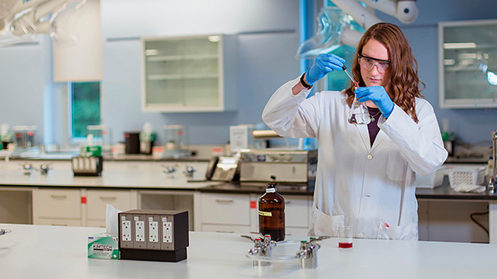 A student in PPE holds a pipette and flask in a chemistry lab