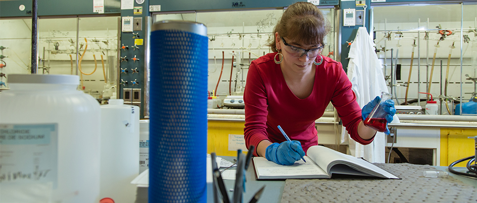 UVic undergraduate student making notes in a book while holding a beaker containing liquid