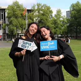 Two students in graduation robes holding signs reading 'super geeky' and 'proud Dad of a UVic grad'