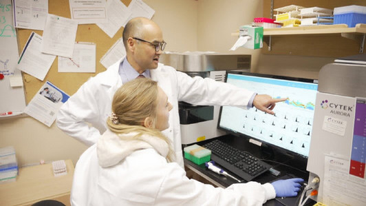 Two scientists in white lab coats looking at computer monitor wiht scientific data displayed.