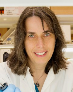 headshot of smiling Dr. Caren Helbing wearing a white lab coat with laboratory shelves in background. 