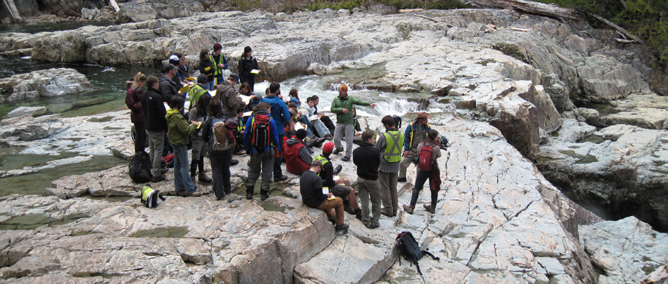 A large group of students standing on rocks outside listening to a professor