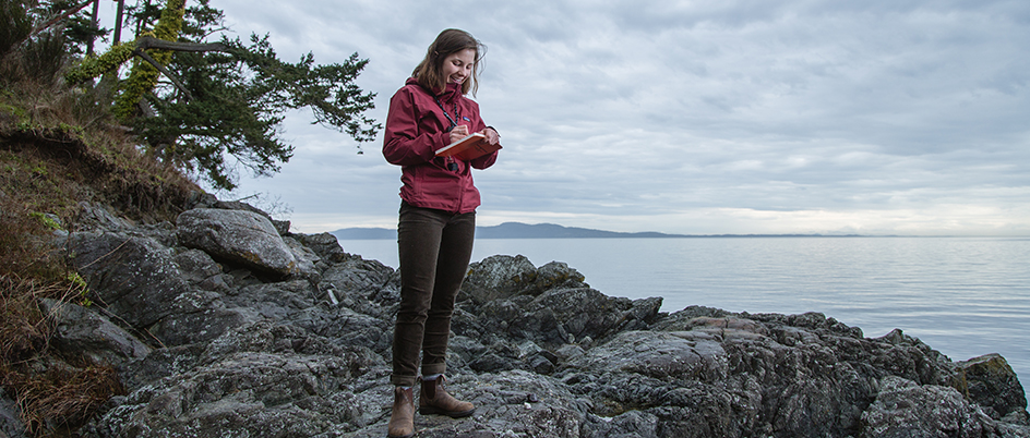 Student standing on rocks near the ocean writing in a notepad