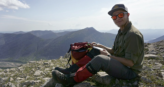 Student sitting on the Mackenzie Mountains with a backpack and notepad