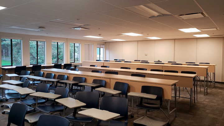 View from the front of the room showing moveable chairs with attached tablet desks and four rows of tall tables with chairs facing the presenter area.