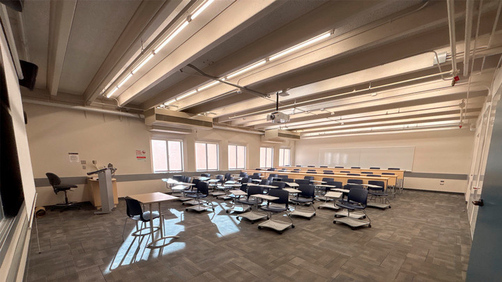 View from the front of the room showing moveable chairs with attached desks with three rows of tall tables and chairs behind them.