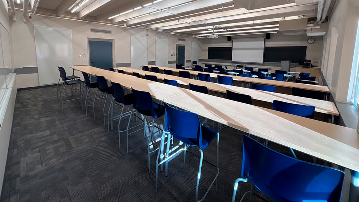 View from the back of the room showing three rows of tall tables with chairs and moveable chairs with attached desks in front of them facing the presenter area.