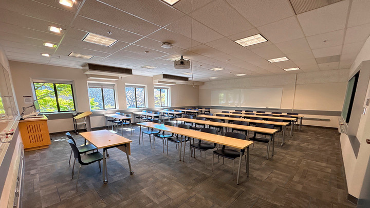 View from the front of the room showing rows of tables and chairs facing the presenter area. The presenter area also has a table and chair, podium and AV cabinet.