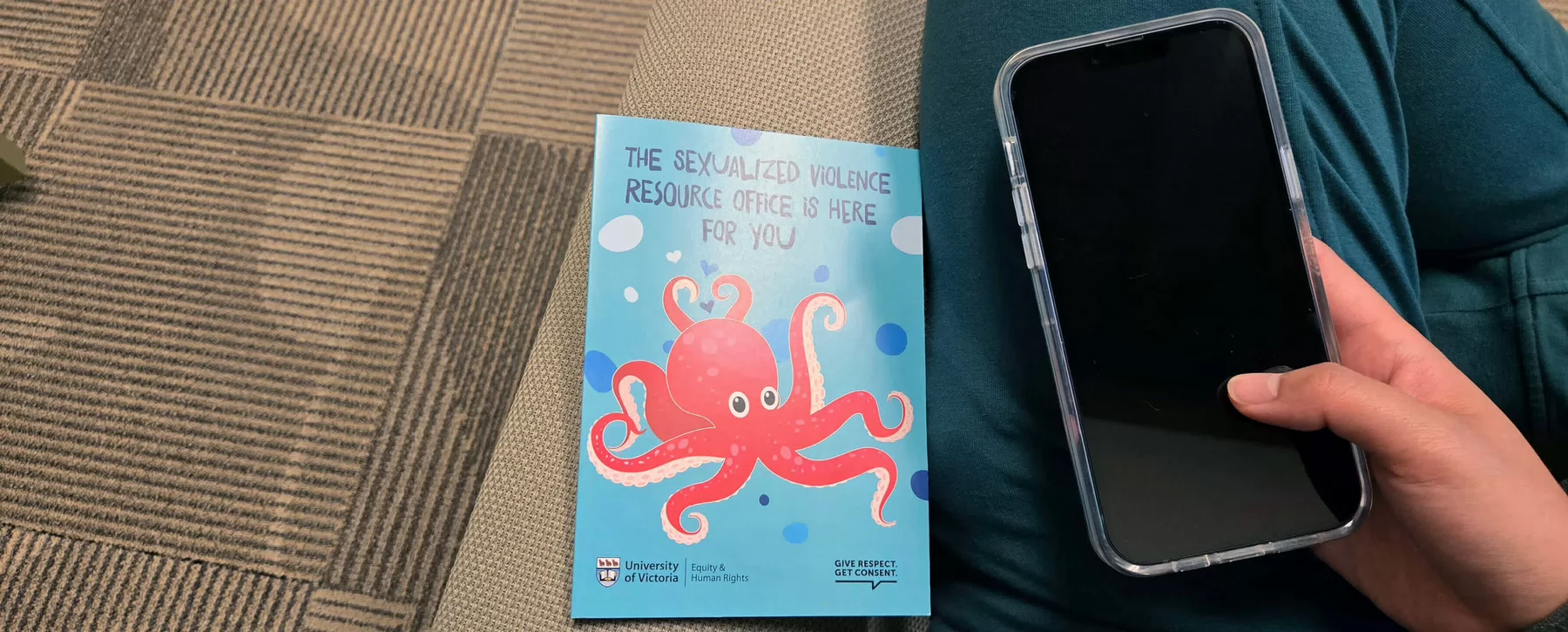 A person holds a smartphone next to a brochure on sexualized violence resources at UVic