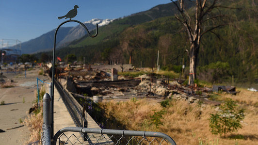 A metal bird d ecoration sits on a fence in front of charred ruins, with green hills and mountains in the background
