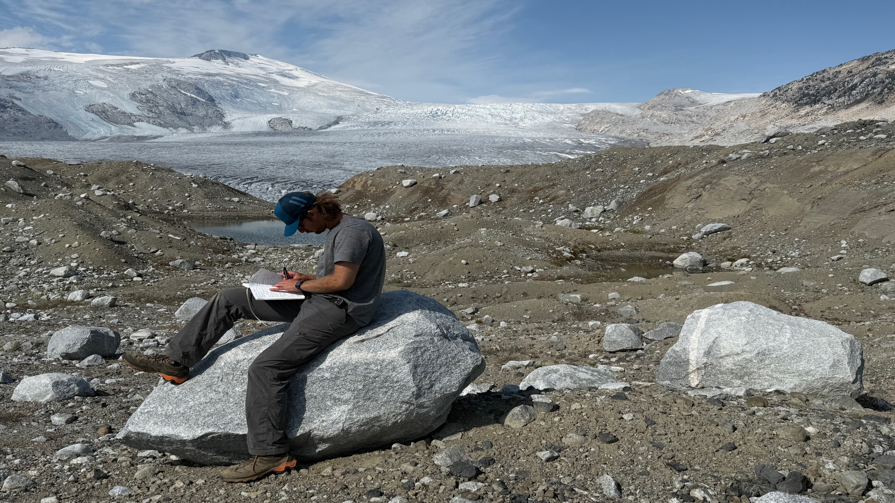 Student sitting on rock making field notes with the mountain range behind him