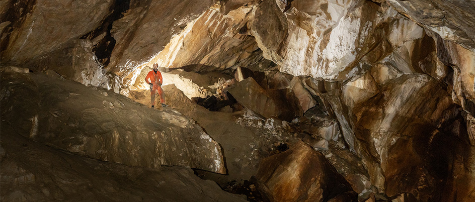A man in an orange jumpsuit and helmet stands in well lit cave